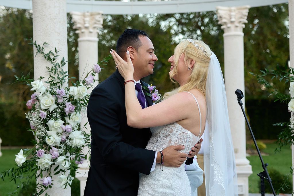 Couple embracing during wedding ceremony, smiling. White dress, black suit, flowers, columns, outdoors.