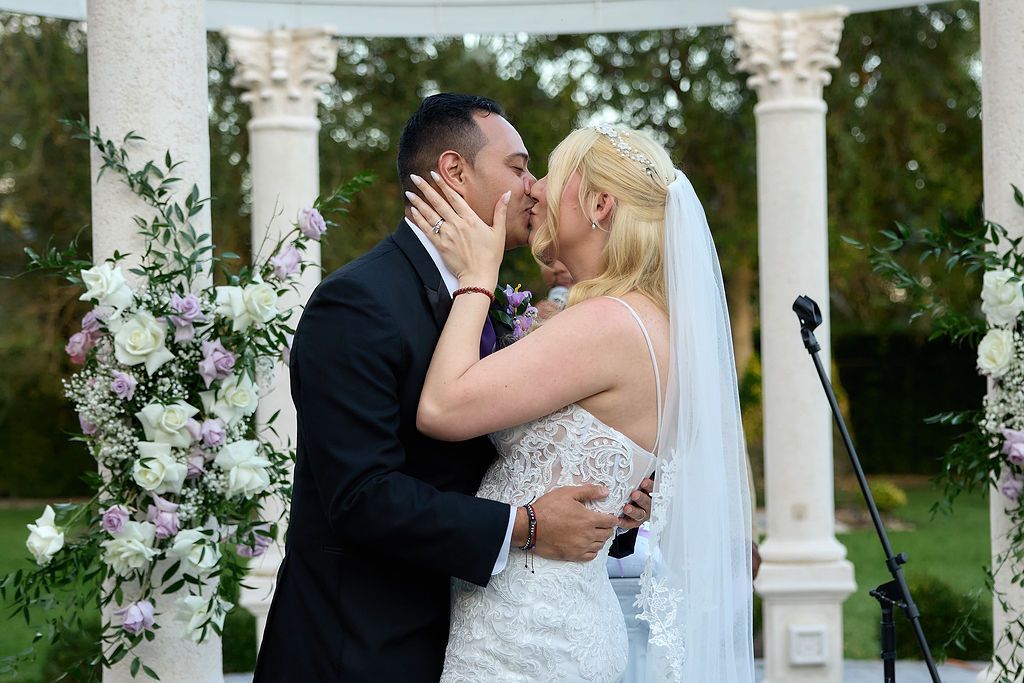 Couple kissing during wedding ceremony under an arch decorated with flowers.