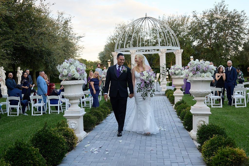 Newlyweds walk down a stone aisle after the ceremony. They are holding hands, with the backdrop of an arbor and floral arrangements.