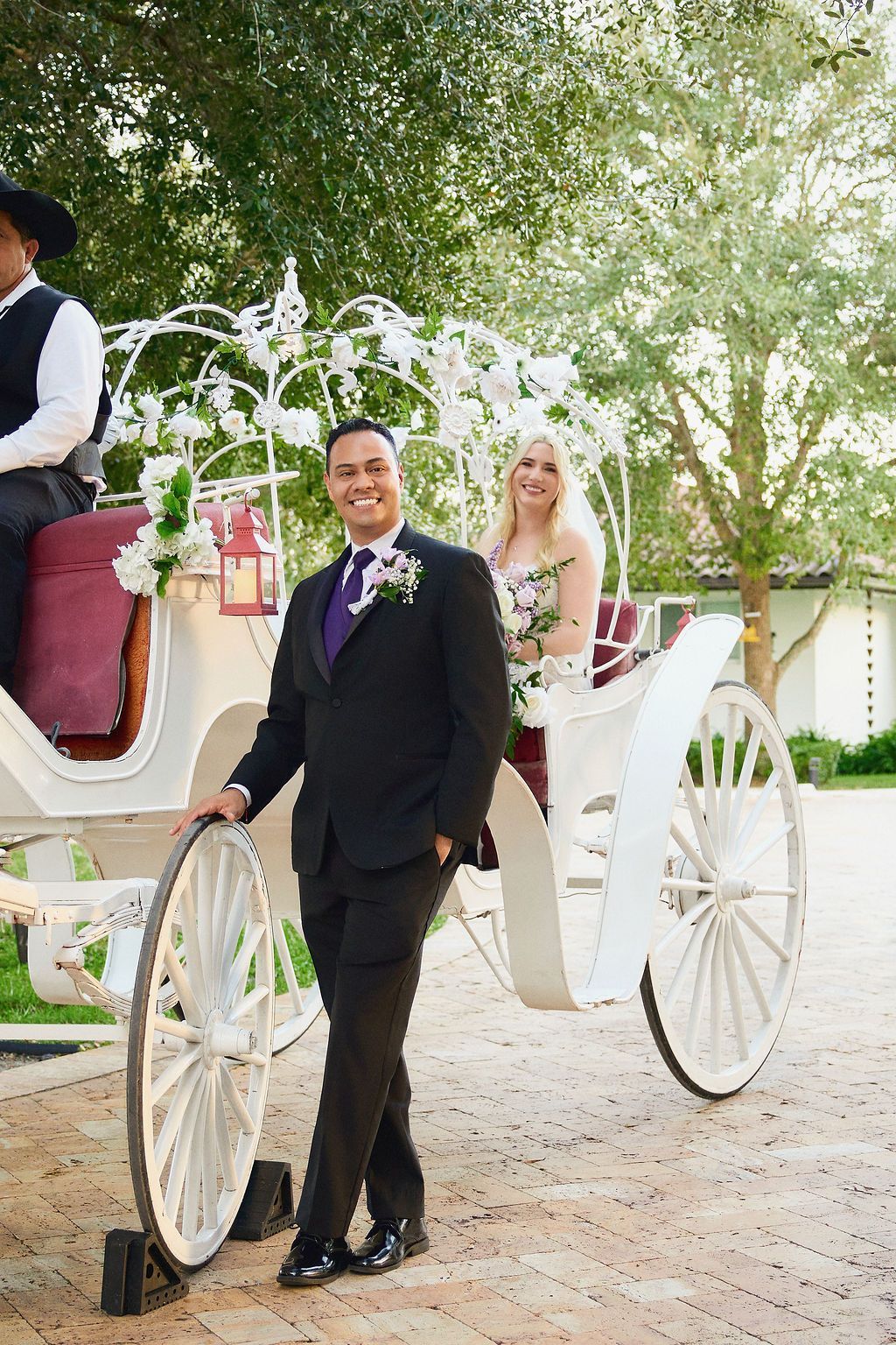 Man in suit stands beside white horse-drawn carriage with bride inside; outdoor setting.