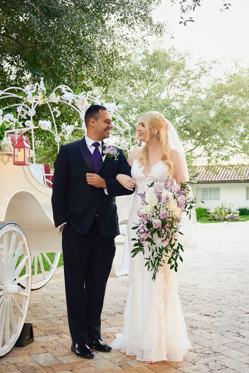 Bride and groom arm-in-arm, smiling, next to a white carriage adorned with flowers; outdoor setting.