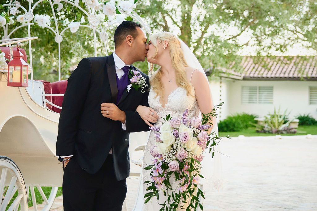 Bride and groom kissing near a horse-drawn carriage; she wears a white dress, he wears a black suit.
