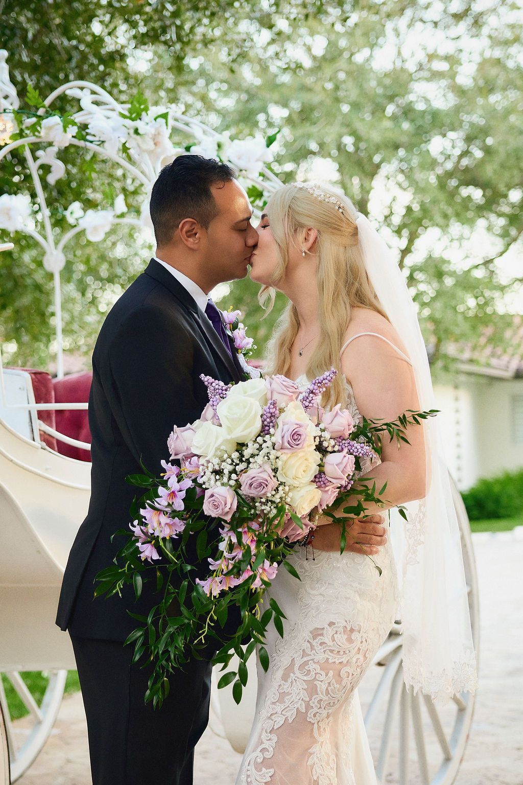 Couple kissing near a white horse-drawn carriage, holding a large flower bouquet.