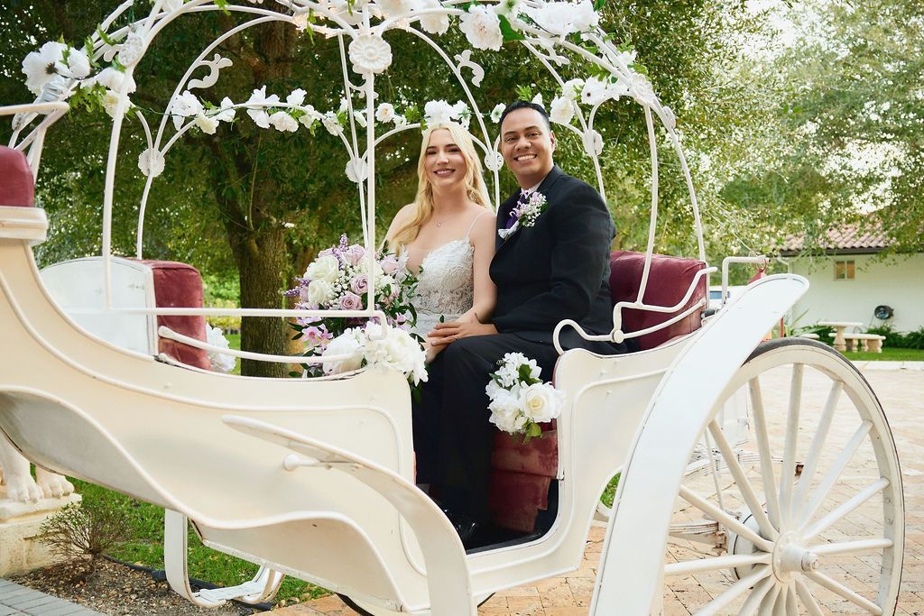 Bride and groom in a white horse-drawn carriage decorated with flowers; smiling in a garden setting.