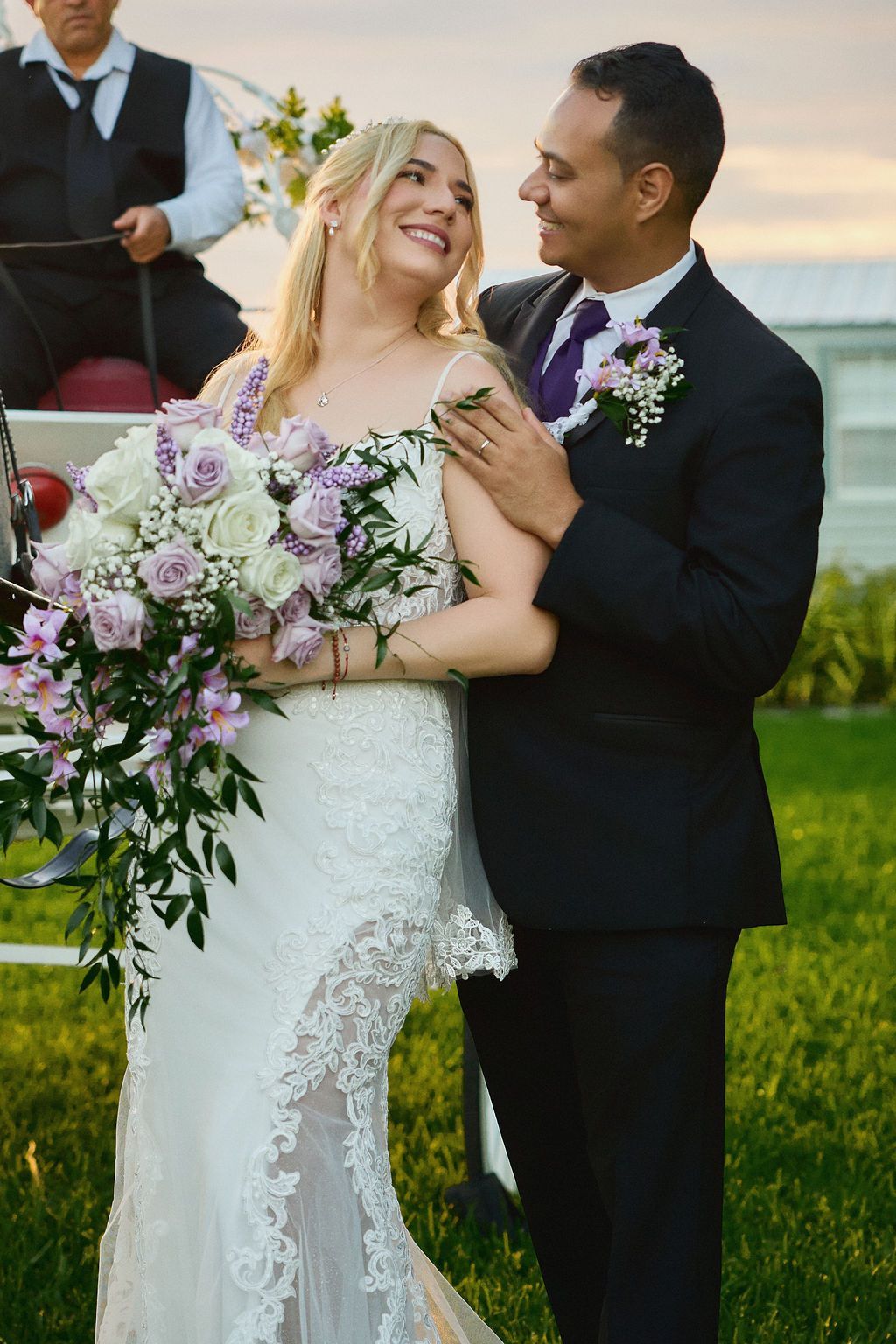 Wedding couple smiles, bride holds flowers. Groom embraces bride, carriage and driver in background.
