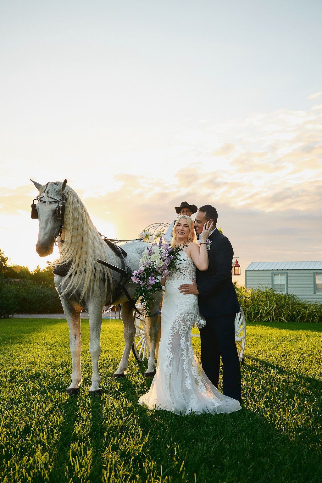 Bride and groom pose with white horse carriage on green lawn during sunset.