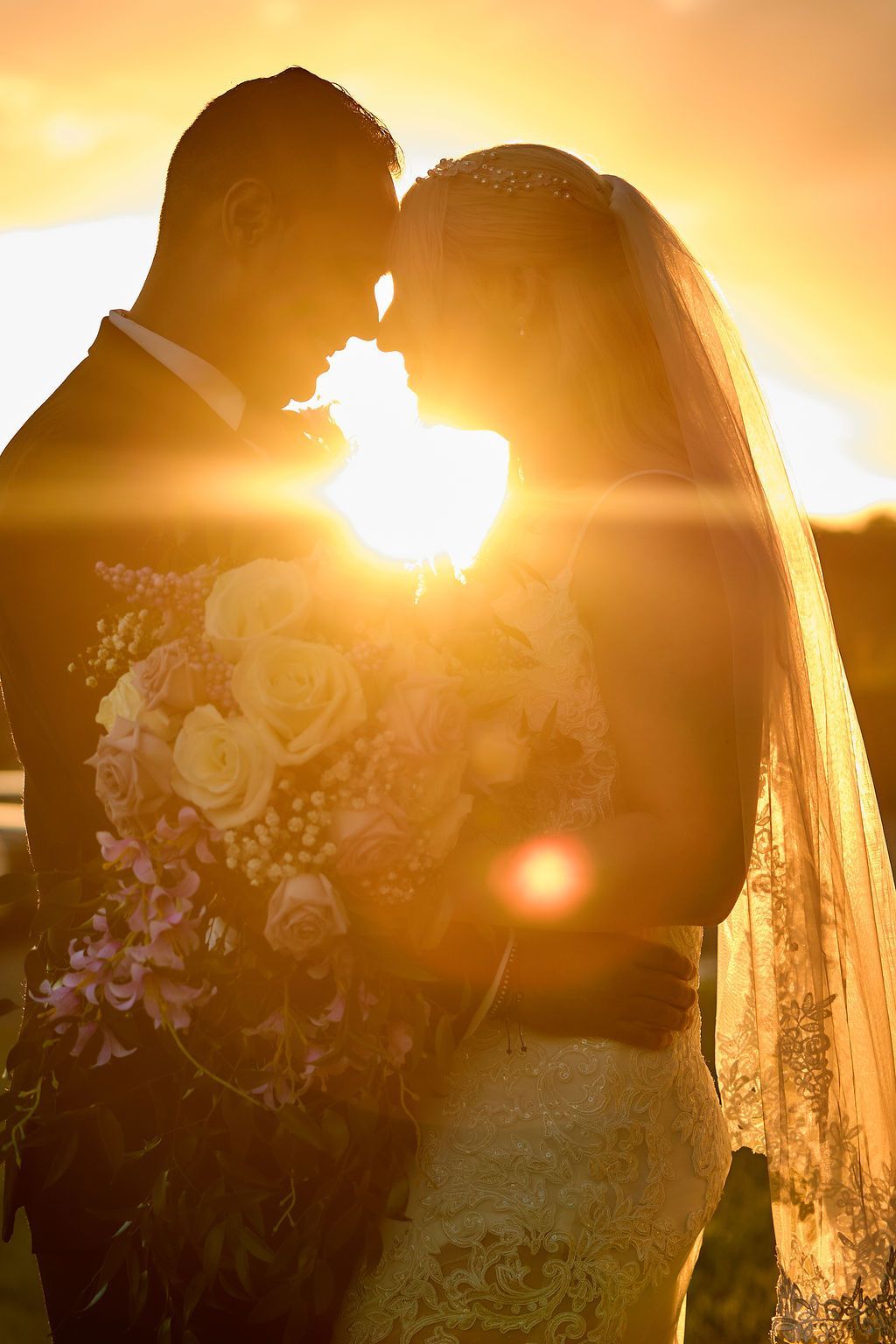 Couple embraces in front of setting sun. Bride holds bouquet, wears veil. Golden light silhouettes their forms.