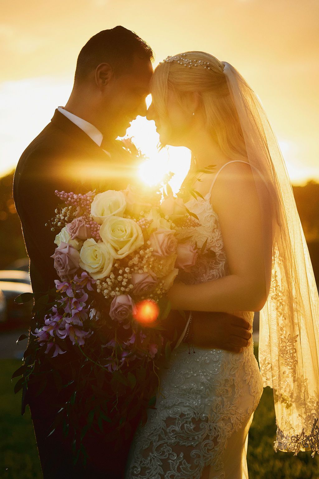 Couple embracing, silhouetted against a golden sunset, bride holding bouquet, wearing veil.