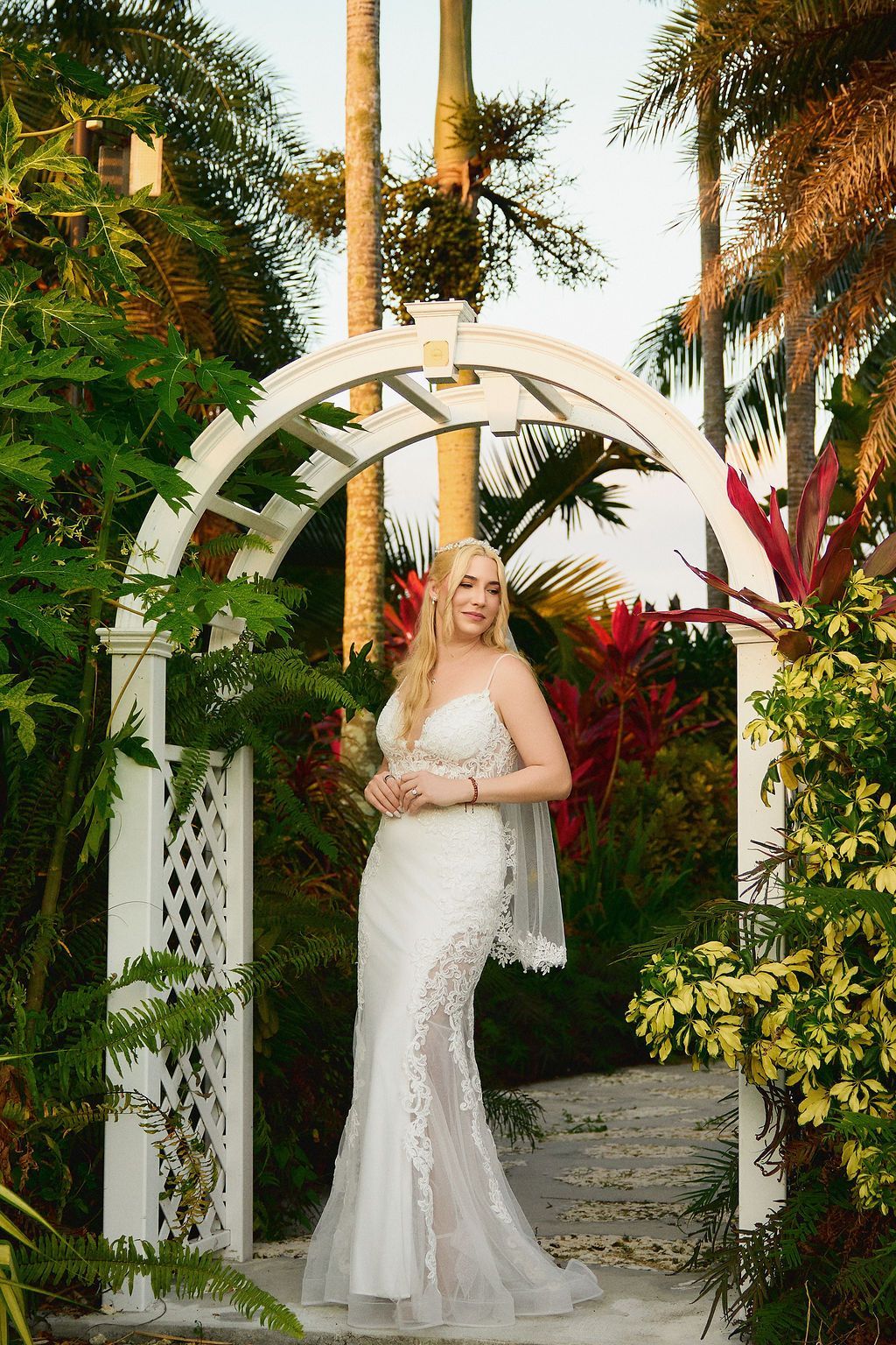 Woman in white wedding dress stands under an arch in a garden, smiling.