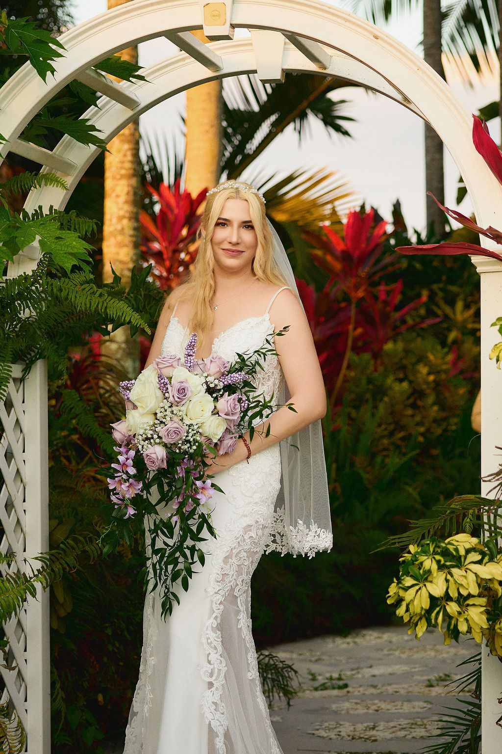 Bride in white lace wedding dress holding bouquet under an arbor.