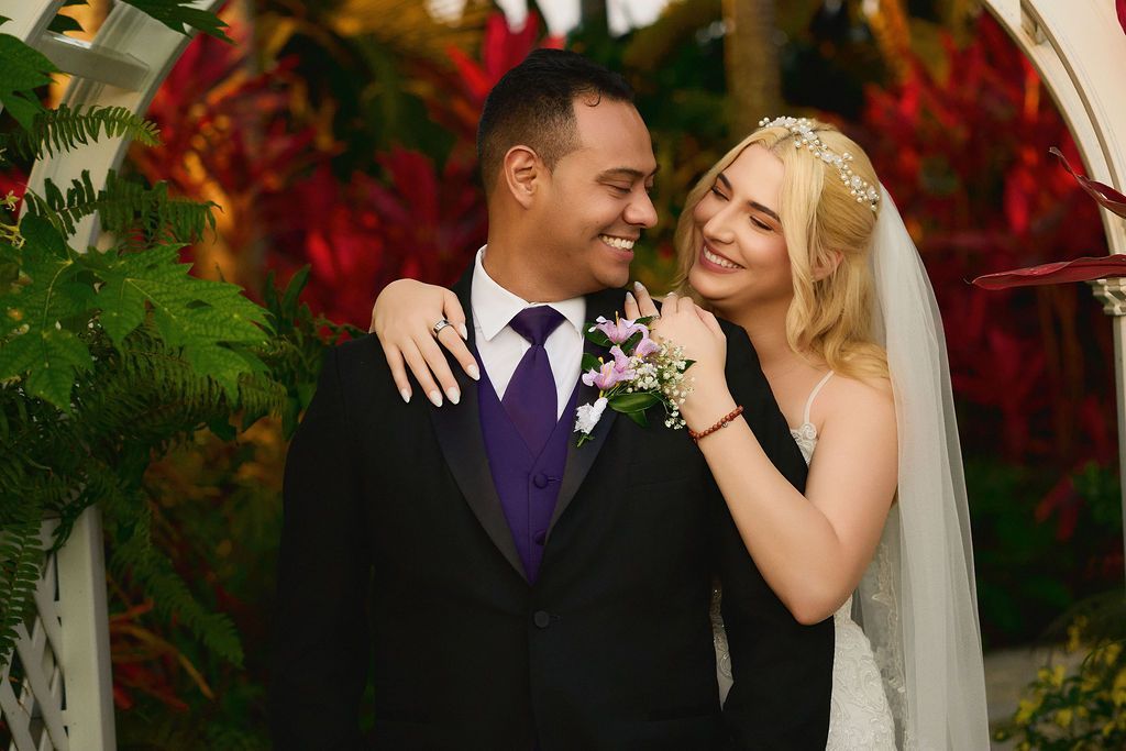 Bride in white dress embraces groom in black suit; smiling, posing outdoors.