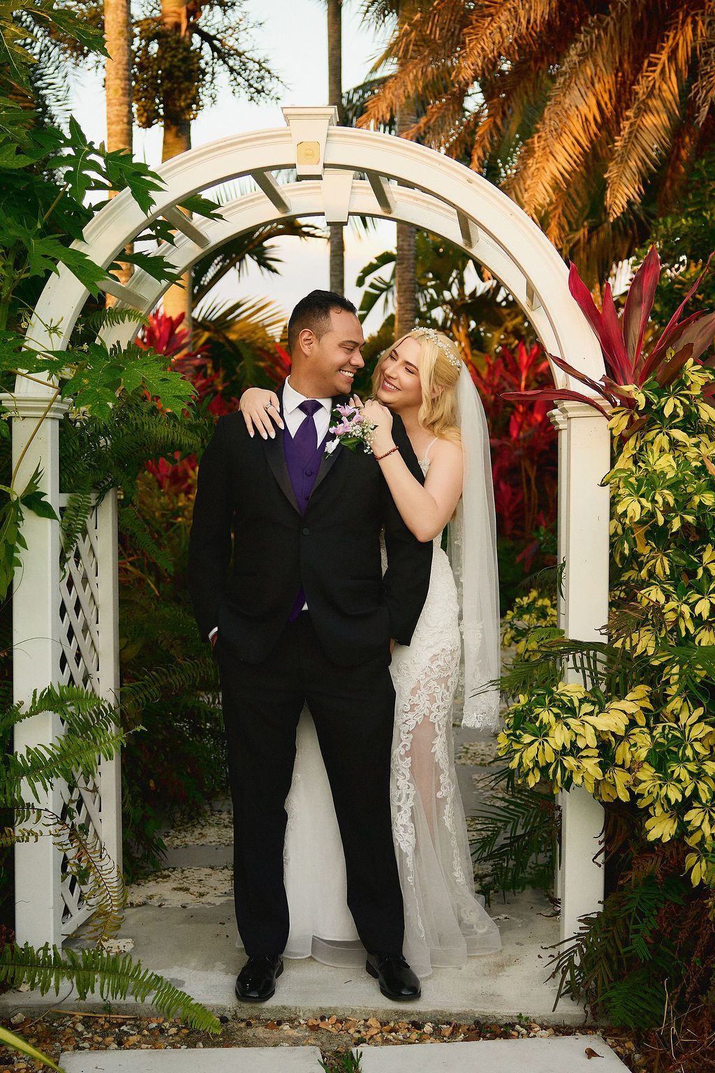 Wedding couple under white archway: Groom in black suit smiles at bride in lace gown; lush garden setting.