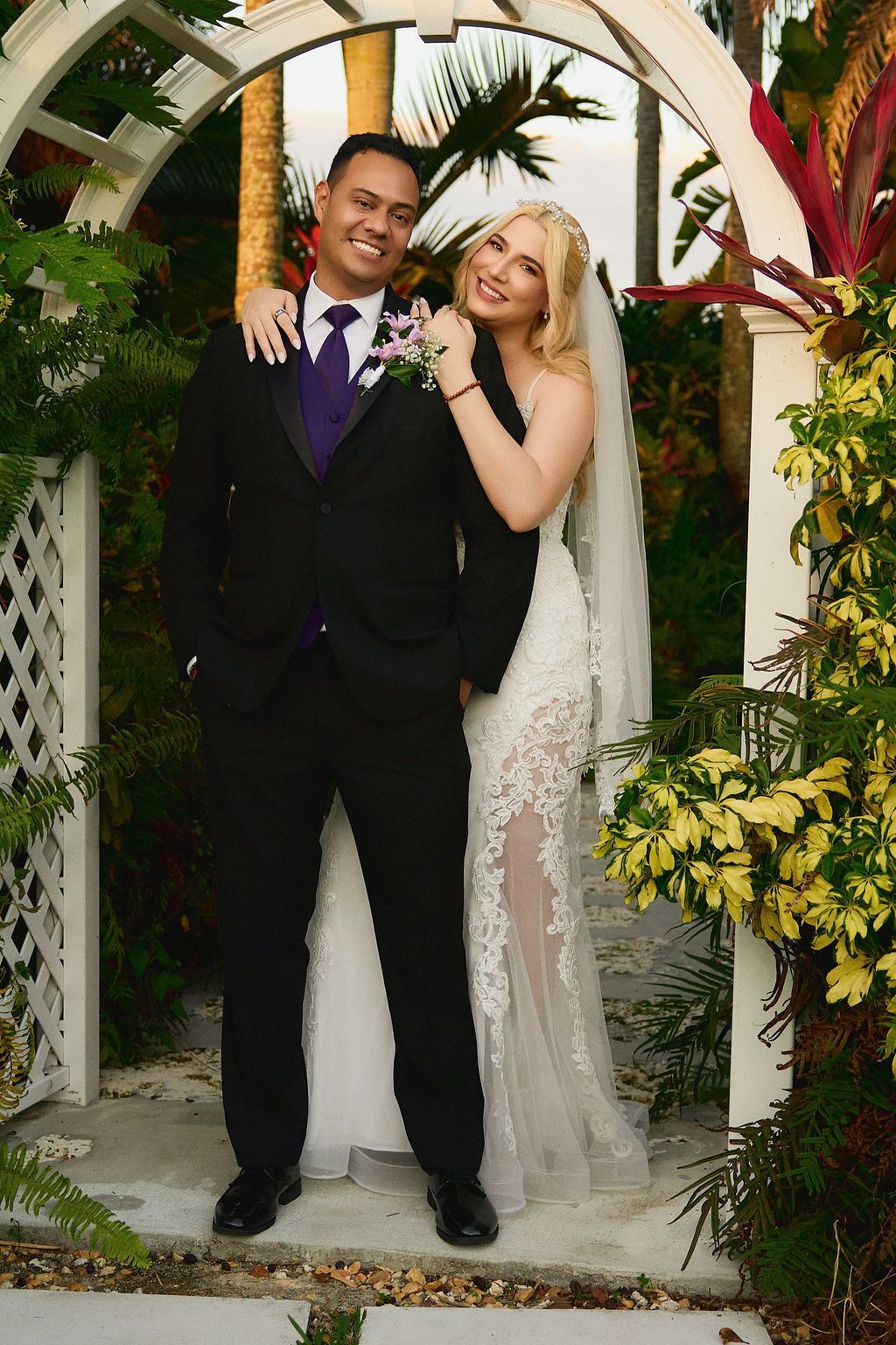 Couple poses under archway, man in black suit, woman in white dress and veil, smiling.