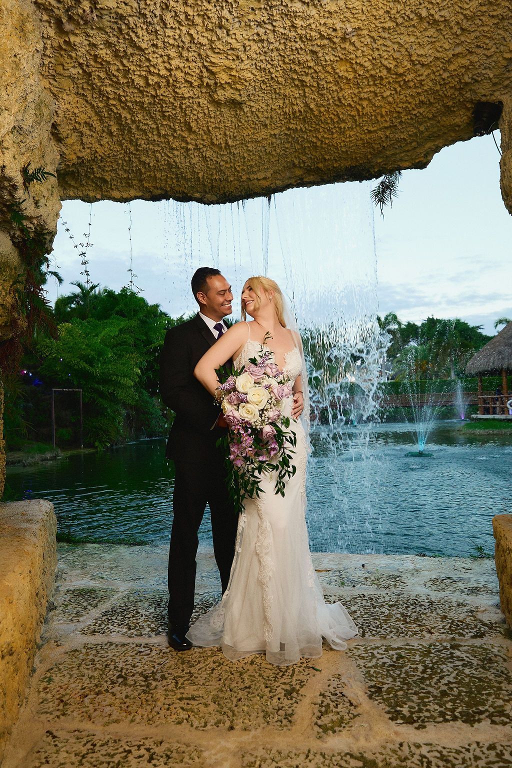 Newlyweds pose under waterfall, holding bouquet, gazing at each other. Black suit, white gown, stone arch.