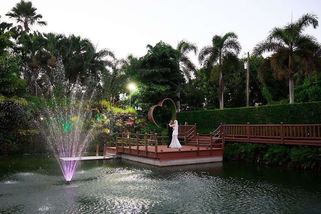Wedding couple on a wooden dock over a pond, fountain in the foreground, palm trees and greenery in the background.