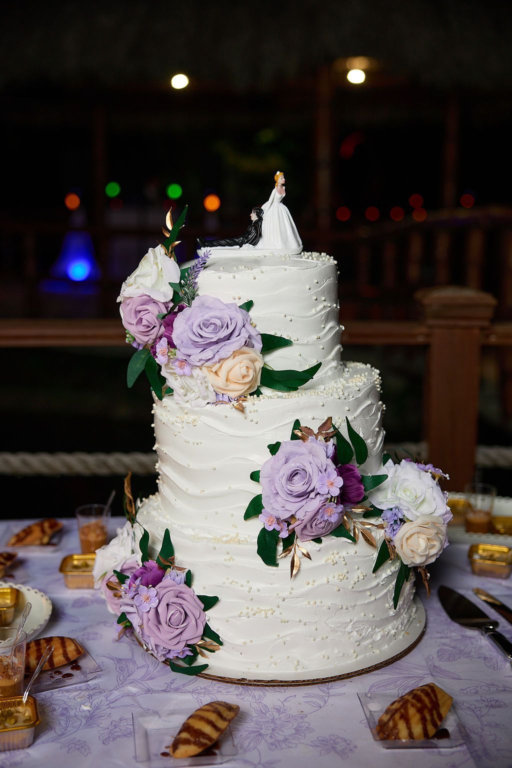 Three-tiered wedding cake with purple flowers and bride-groom topper, on a table.
