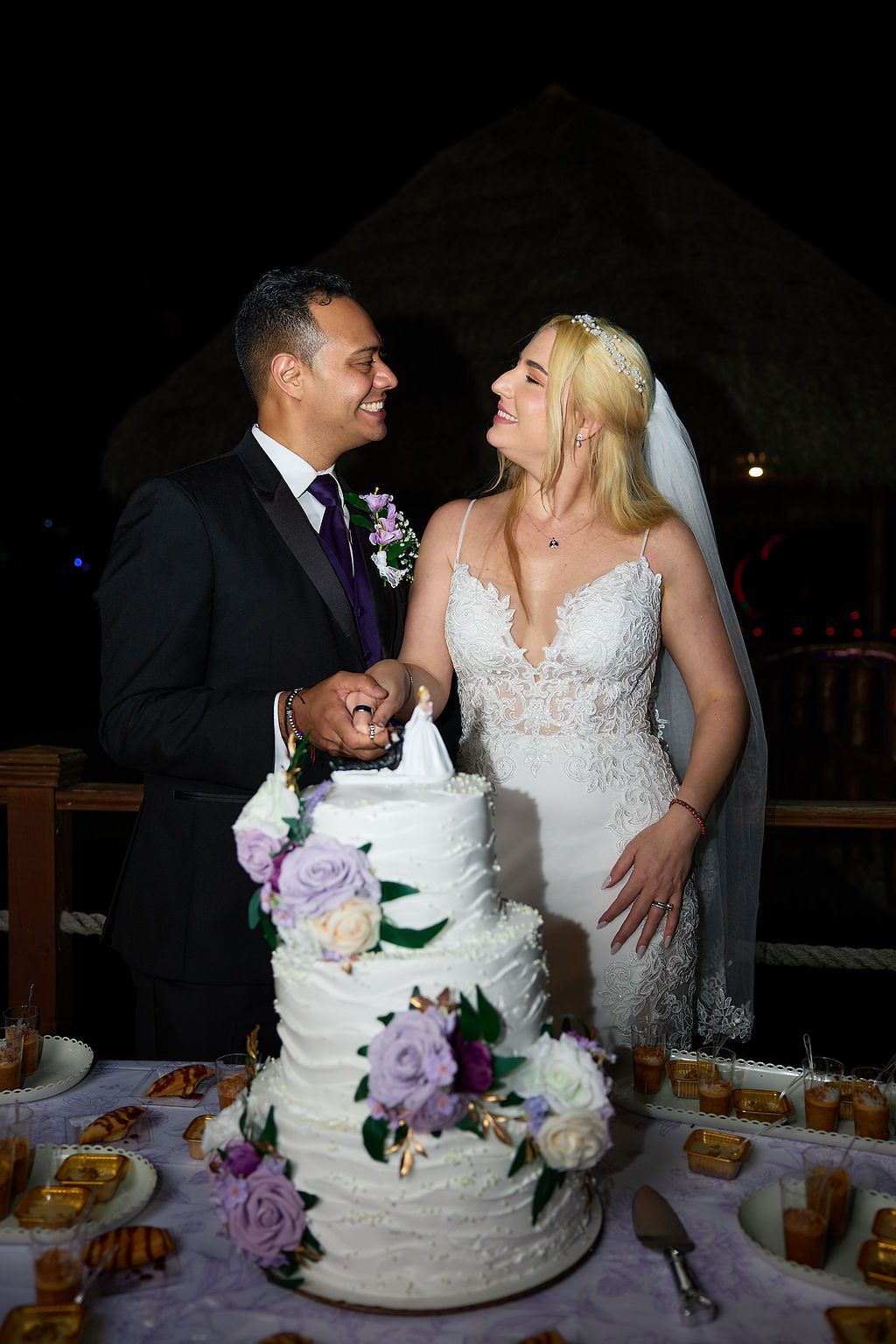 Bride and groom cutting wedding cake at outdoor reception, smiling at each other. Cake has floral decorations.