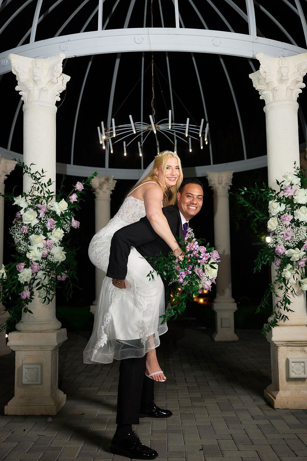 Groom giving bride a piggyback ride under a gazebo decorated with flowers. Smiling, they are lit by a chandelier.