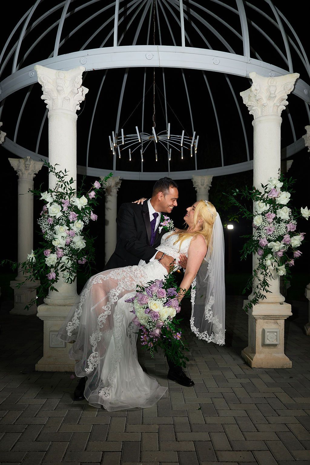 Bride and groom embrace beneath gazebo, surrounded by flowers, at night. She's in a white dress, he wears a suit.