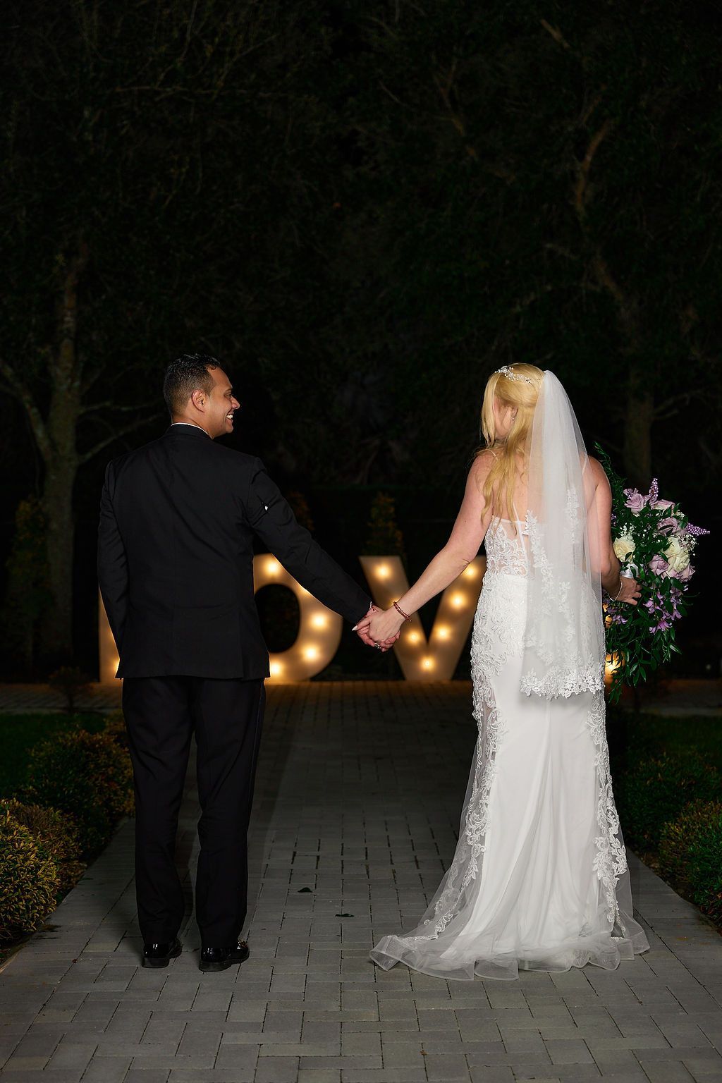 Bride and groom holding hands, walking on a brick path at night toward lit letters