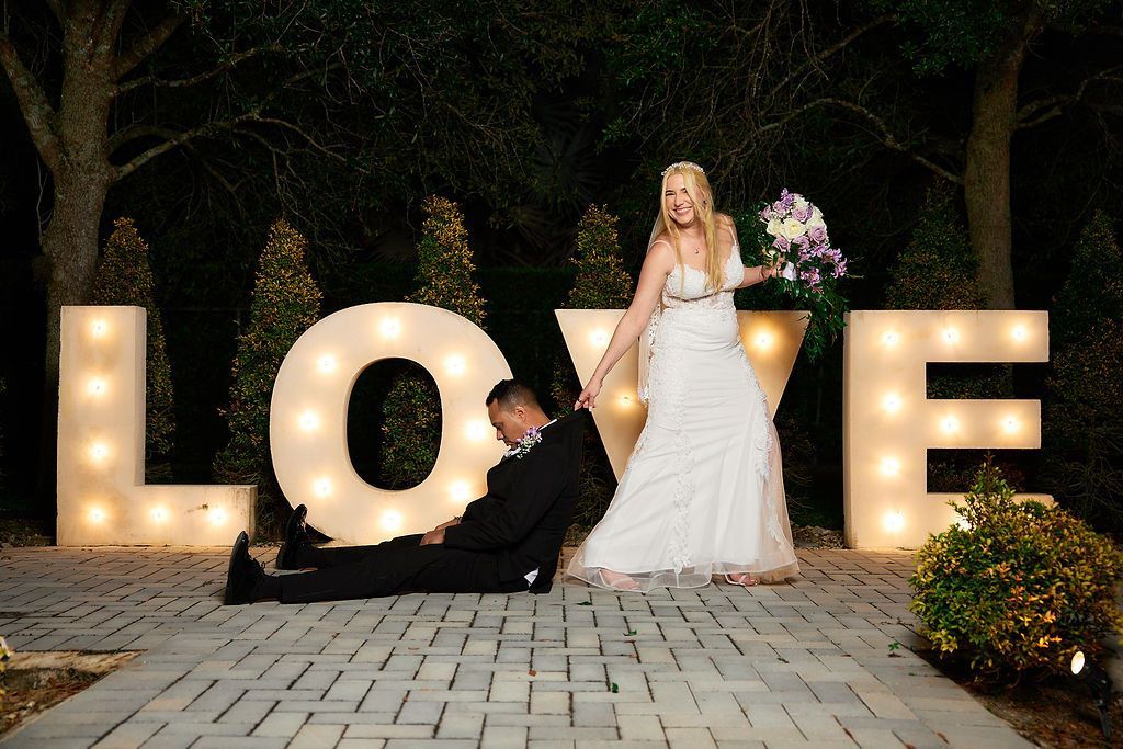 Bride in white dress pulls groom, sprawled on the ground, near illuminated