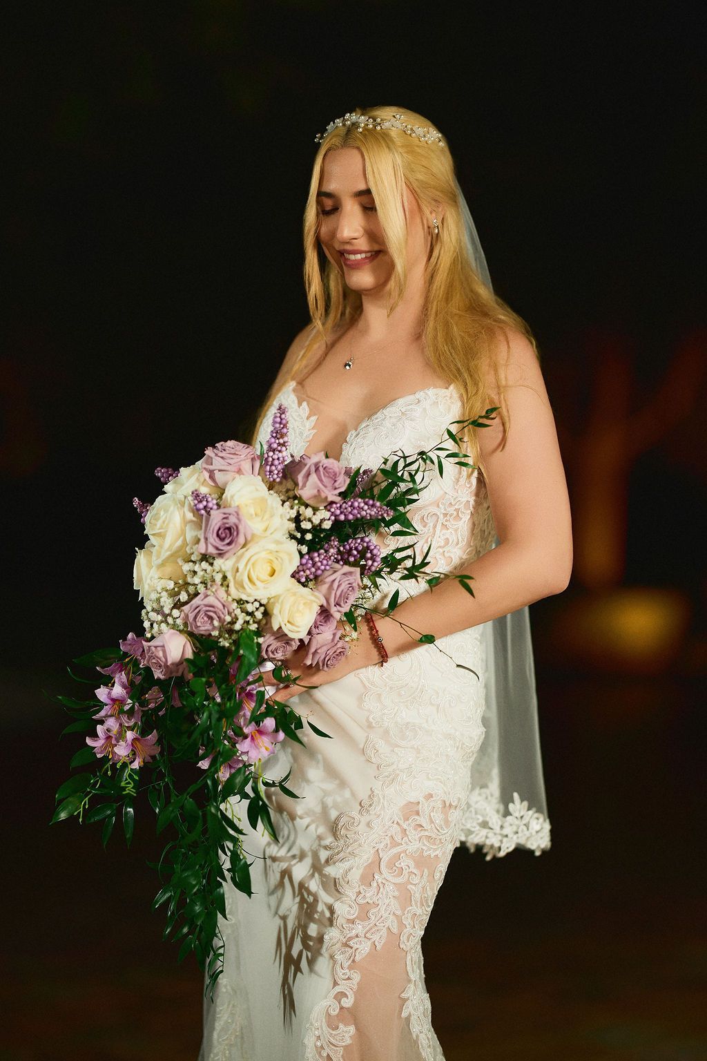 Bride in a white lace wedding dress, holding a bouquet of white and purple flowers, smiling.