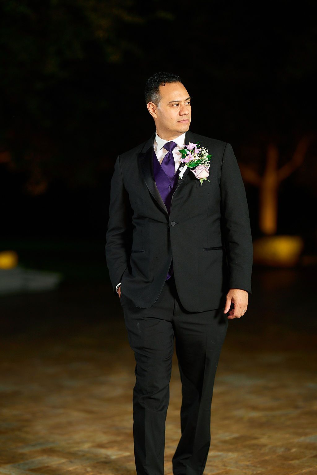 Man in black tuxedo with purple tie and boutonniere, lit by a soft light outdoors.