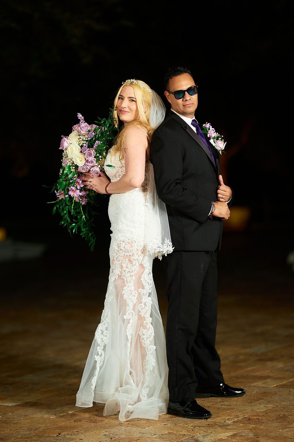 Bride and groom back-to-back, posing for a photo. The bride in a white gown holding flowers, the groom in a suit and sunglasses.