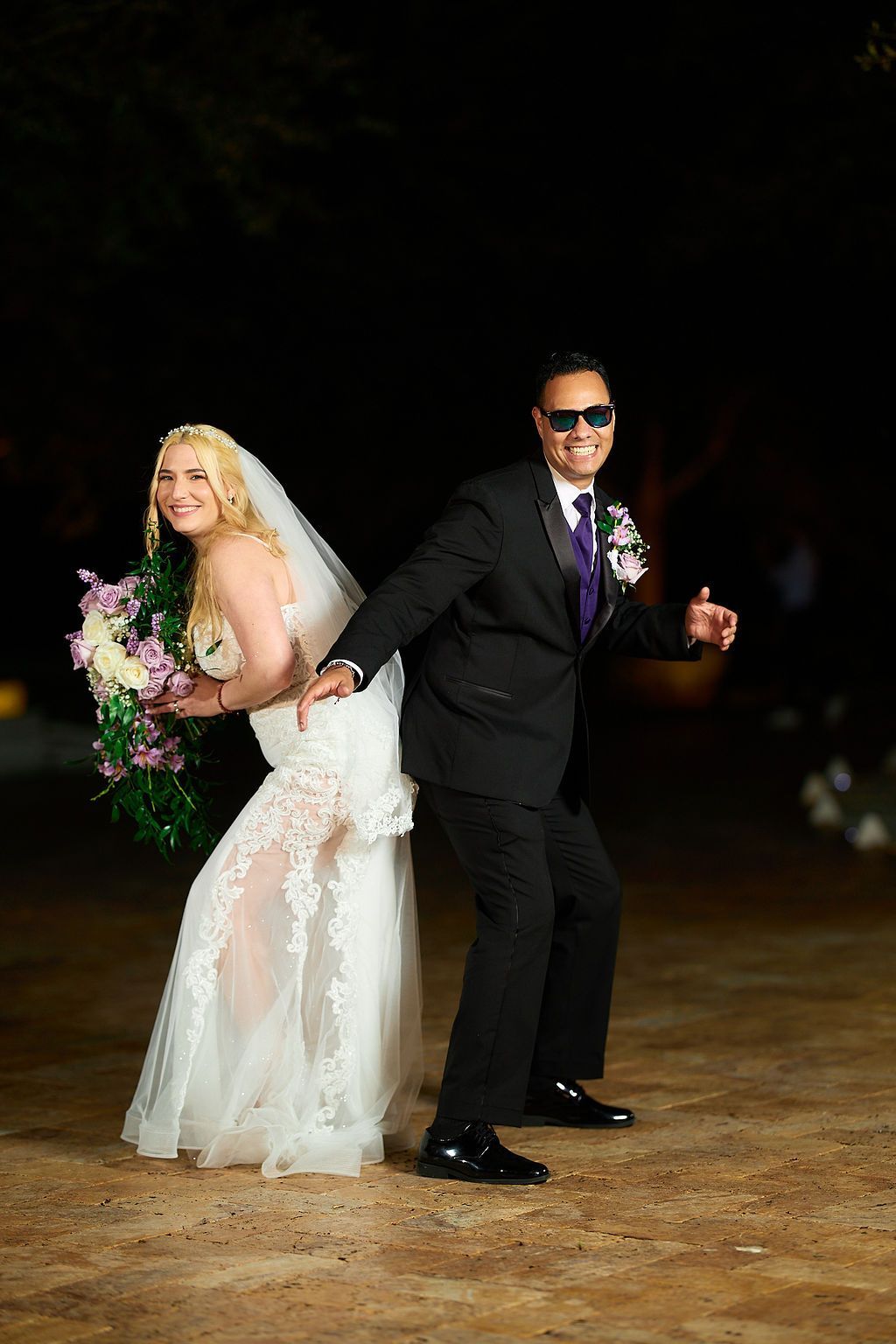 Bride and groom pose back-to-back, smiling outdoors at night. The man wears sunglasses and a tuxedo, while the woman wears a wedding dress.