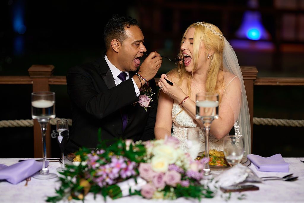 Bride and groom feed each other cake, laughing at a wedding reception table with flowers.