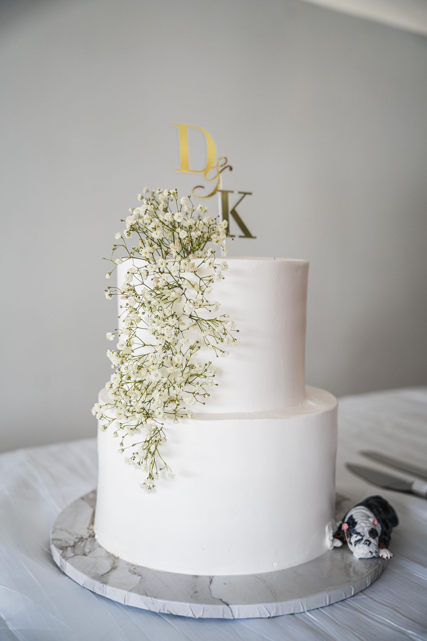 Two-tiered white wedding cake with floral cascade and initials topper, marble base, miniature dog figurine.