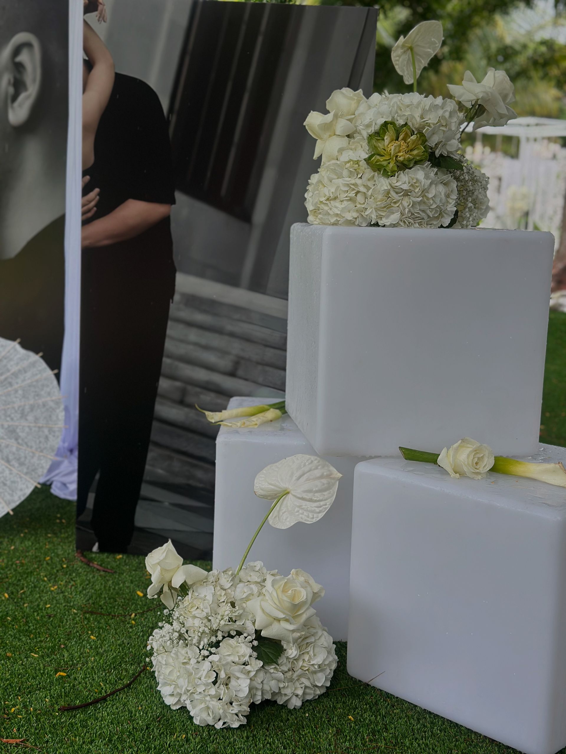 White floral arrangements on stacked white cubes, in front of a photograph and green lawn.