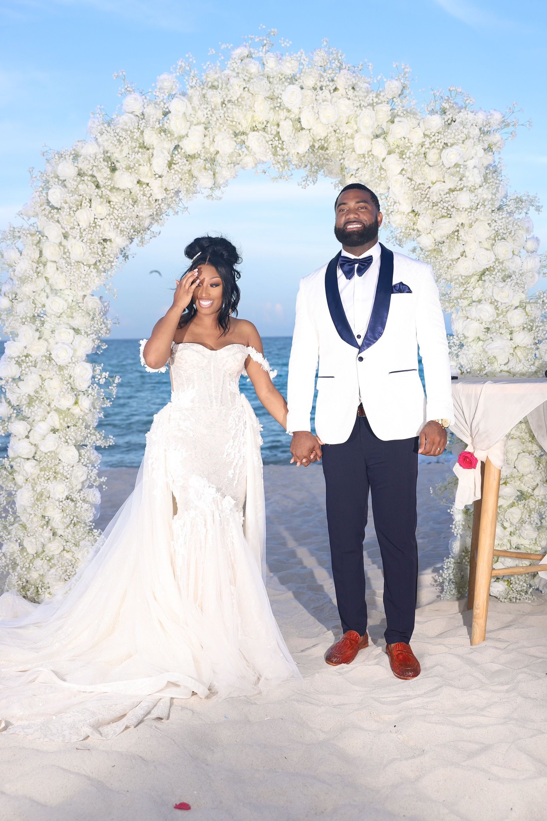 A bride and groom are standing in front of a floral arch on the beach.