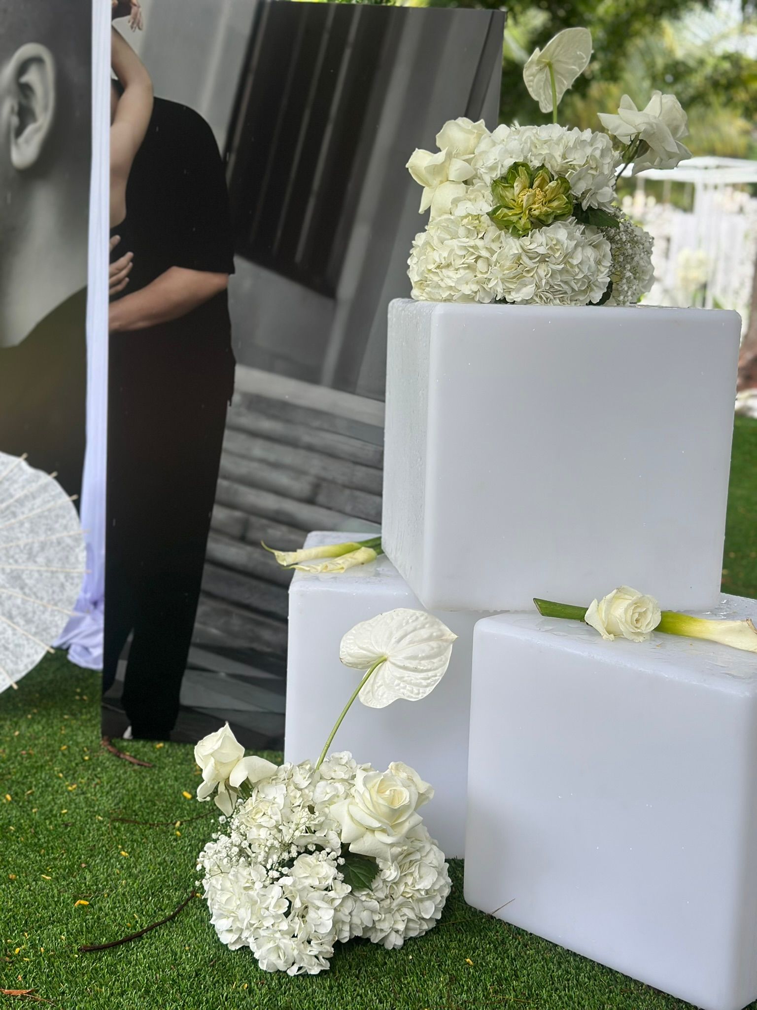 Floral arrangements on white blocks, next to a black and white photo of a person, on green grass.