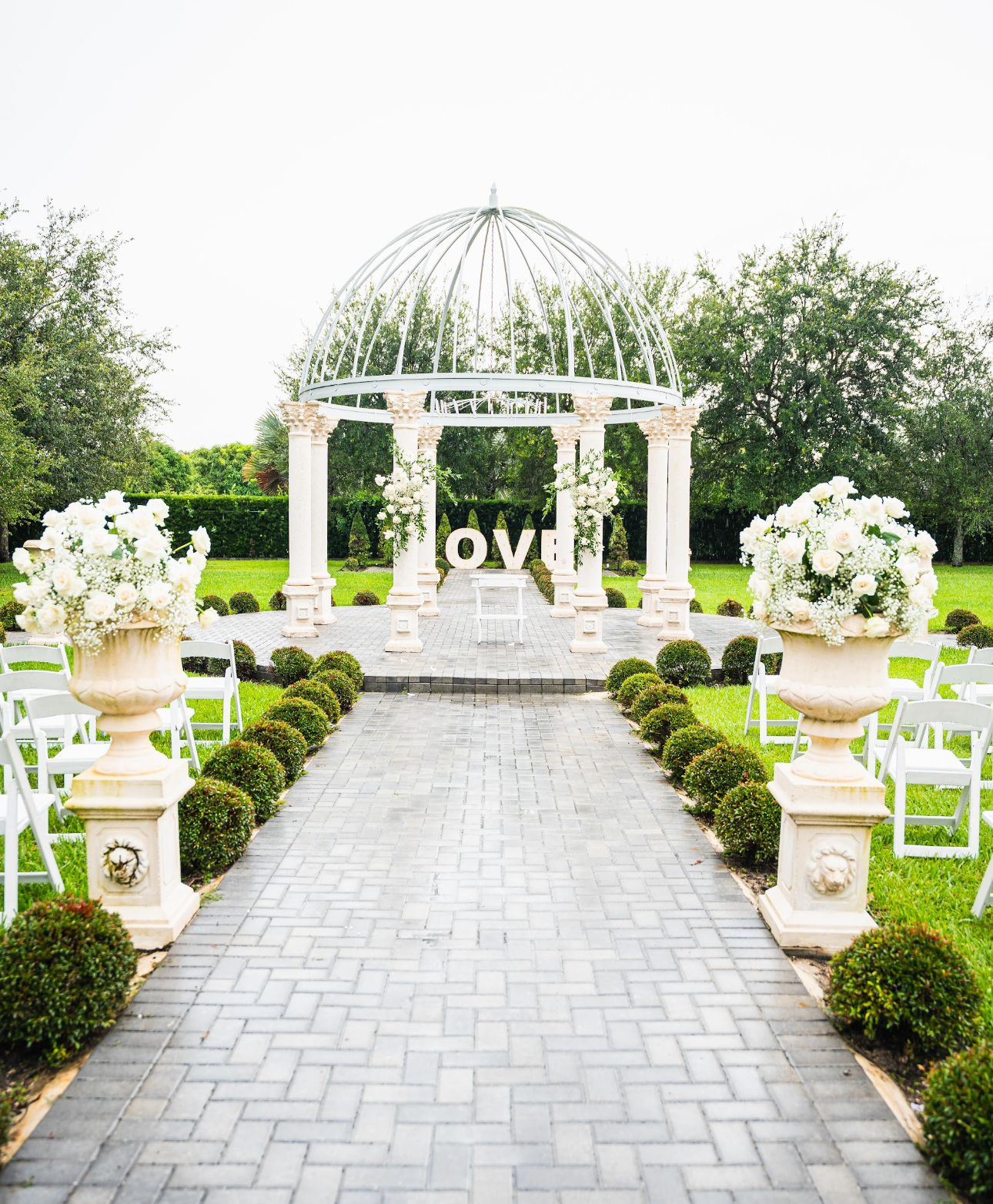 Wedding ceremony setup: Gazebo, pathway lined with greenery, white chairs, floral arrangements.