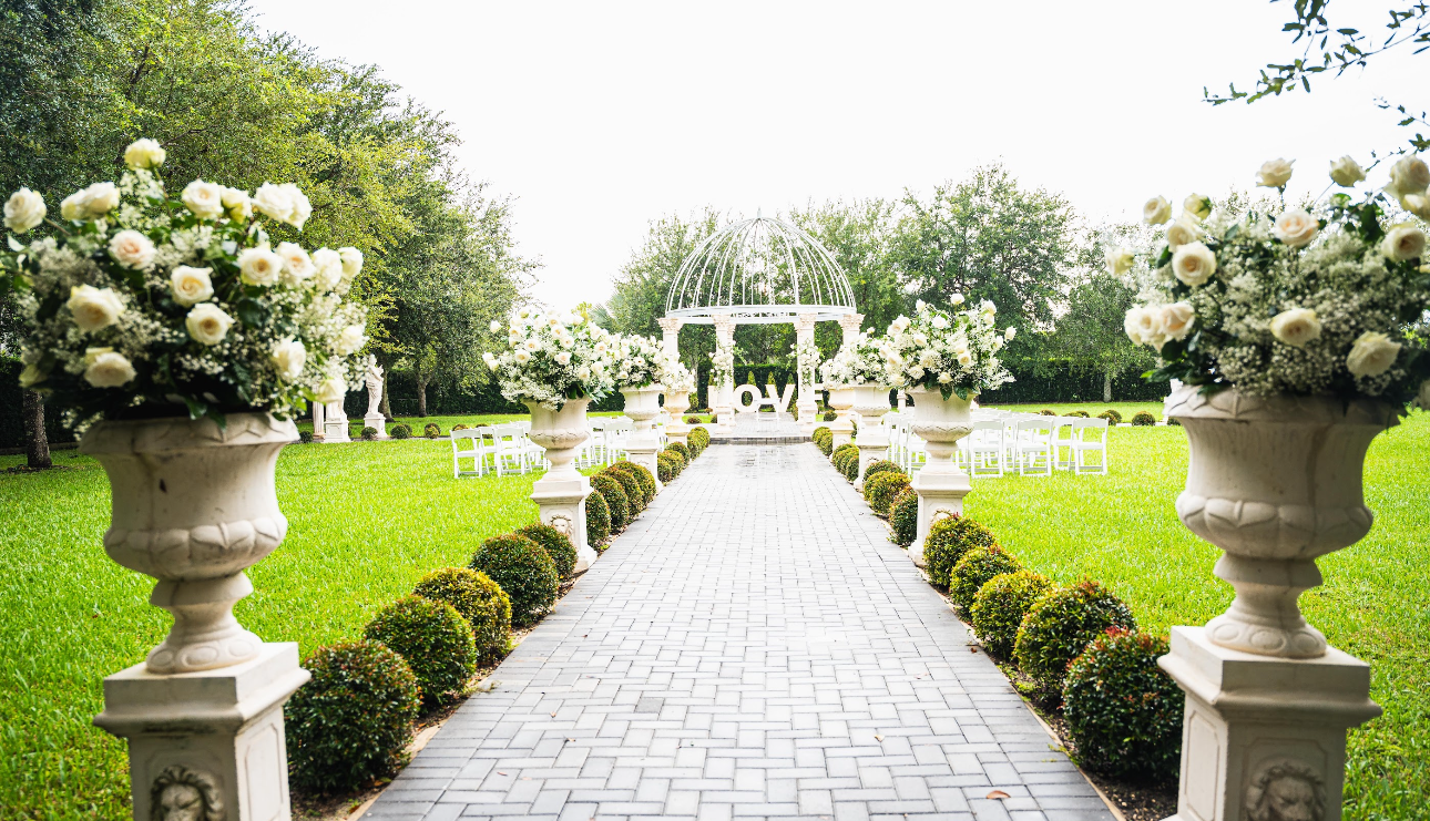 Brick path lined with flower-filled urns leading to a wedding ceremony with a white arbor and green lawn.