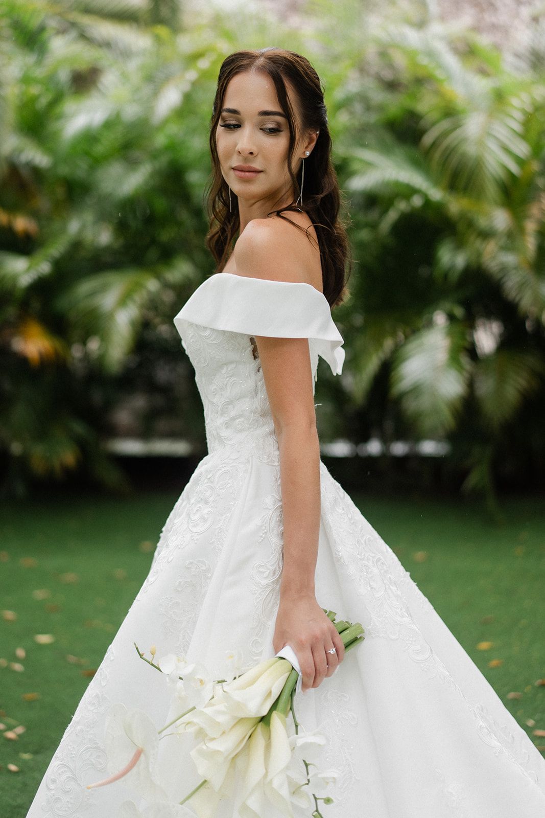 Bride in white off-the-shoulder gown, holding flowers, standing on grass, surrounded by greenery.