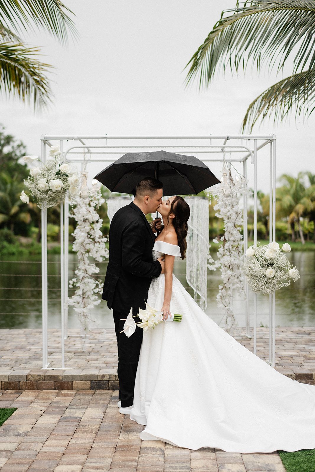 Couple kissing under a black umbrella at their wedding. They stand near a floral arch by a lake.