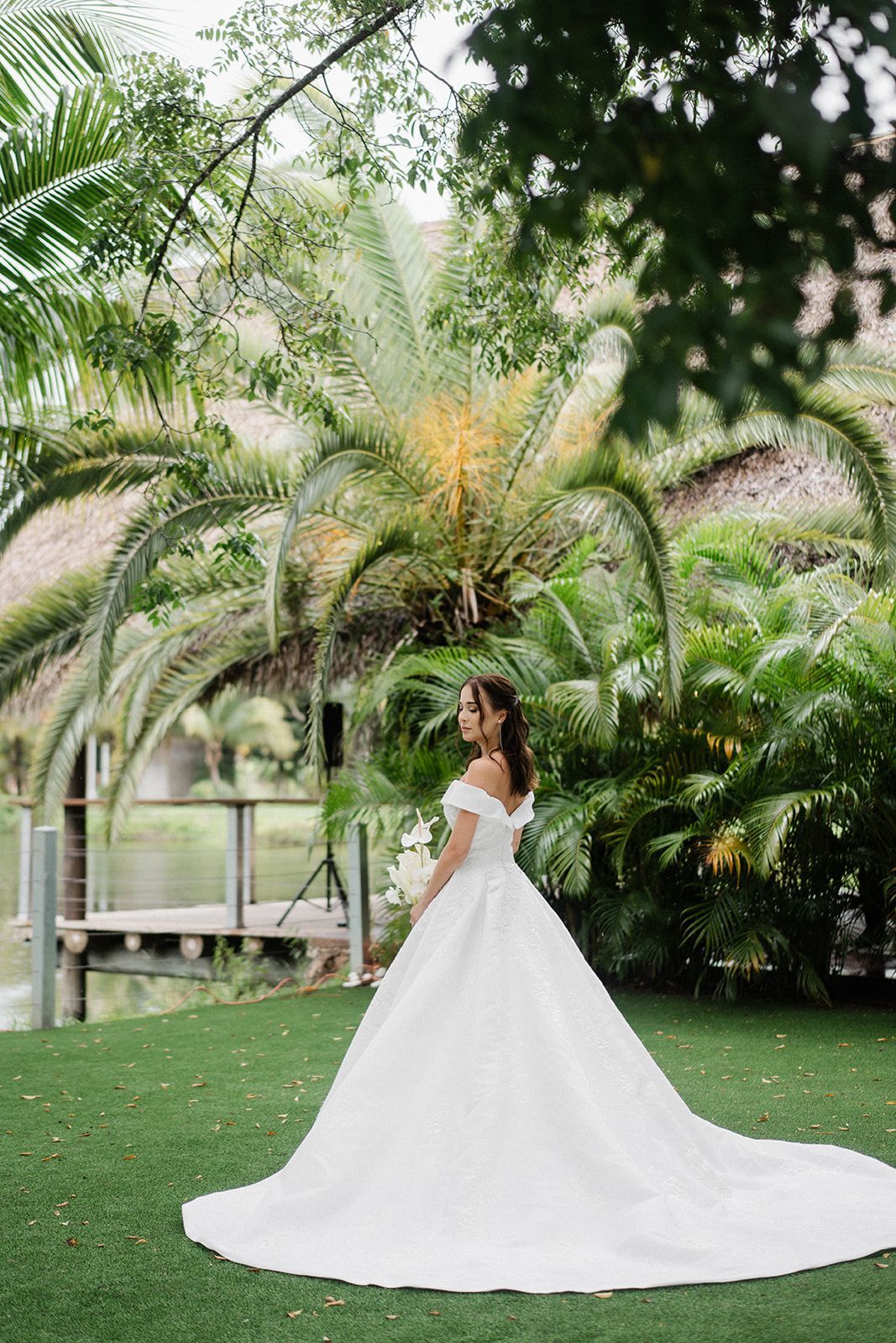 Bride in white dress holding bouquet, posing in front of lush tropical greenery.