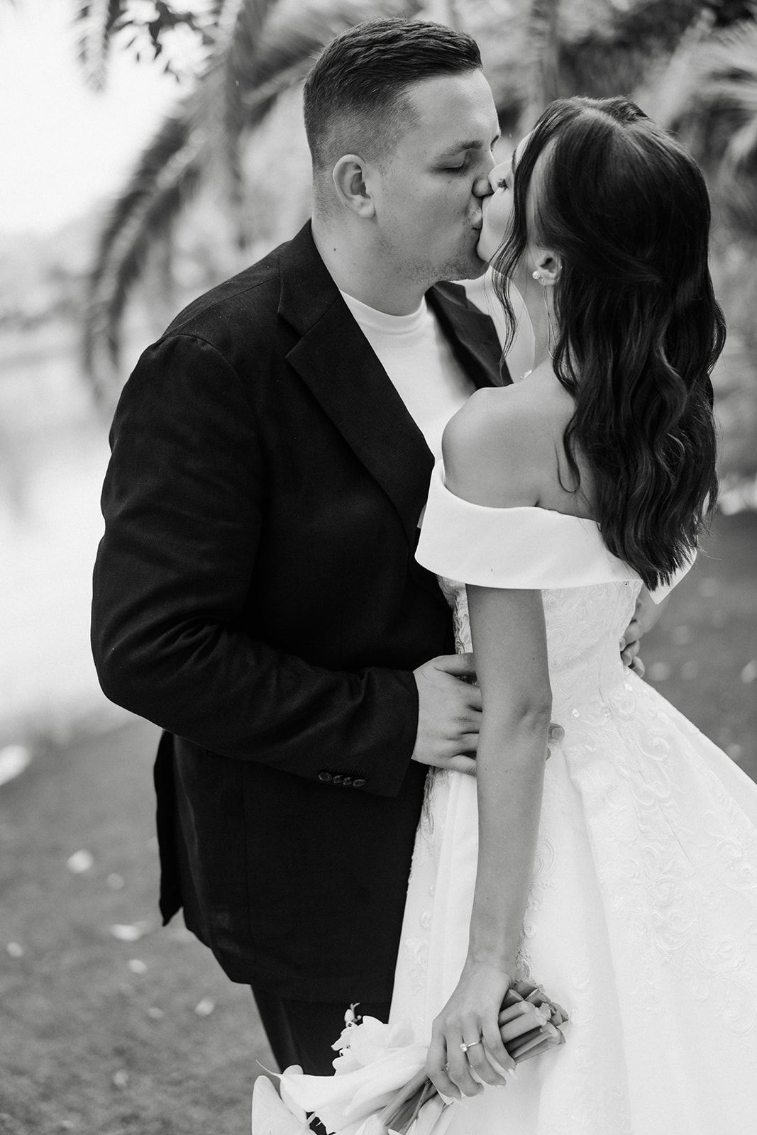 Couple kissing, the woman in a white off-shoulder gown, the man in a black blazer, outside by a body of water.