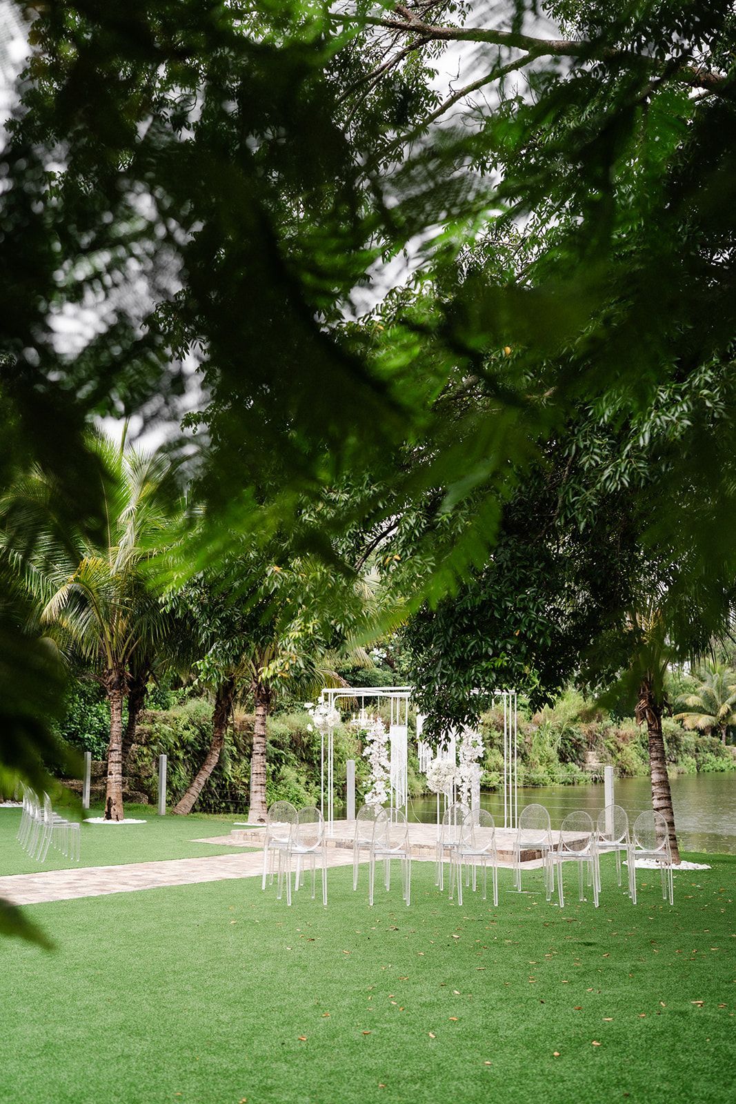 Outdoor wedding setup on a green lawn with transparent chairs and a white arch, framed by trees.