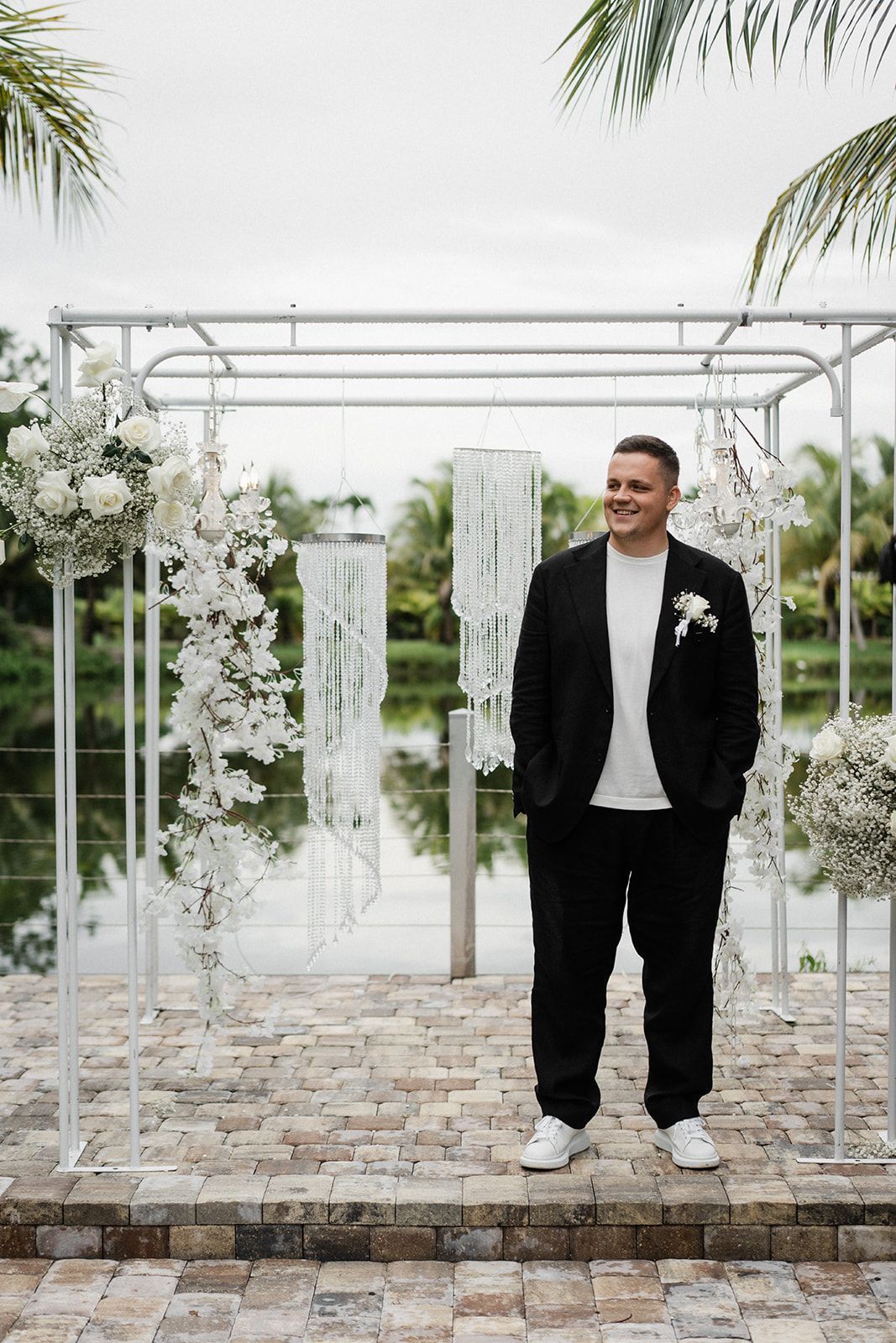 Man in black suit and white sneakers stands by floral wedding arch, smiling, near water.