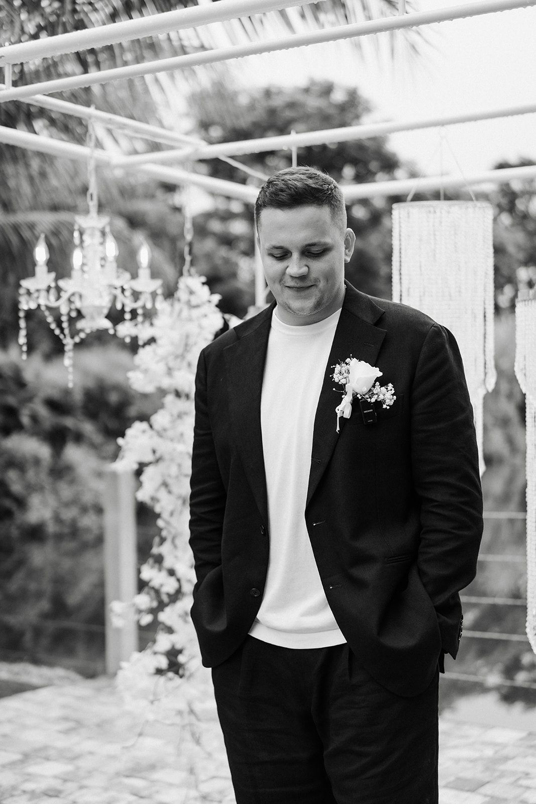 Man in black suit and white shirt, outdoors with floral decorations, looking down.