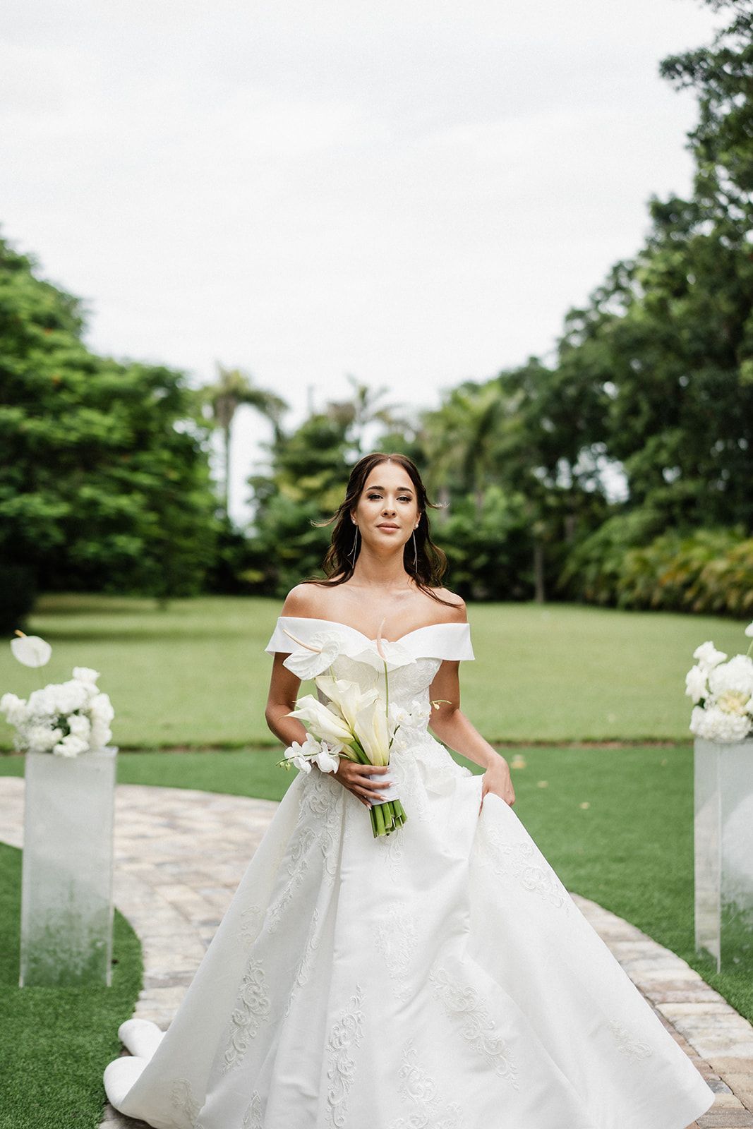 Bride in white gown holding bouquet, walking on stone path, outdoor garden setting.