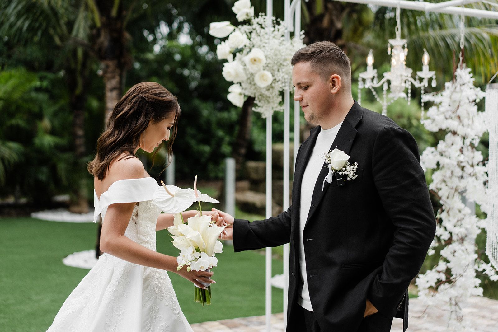 Bride and groom at outdoor wedding ceremony; bride in white dress, groom in suit, holding hands near white floral arch.
