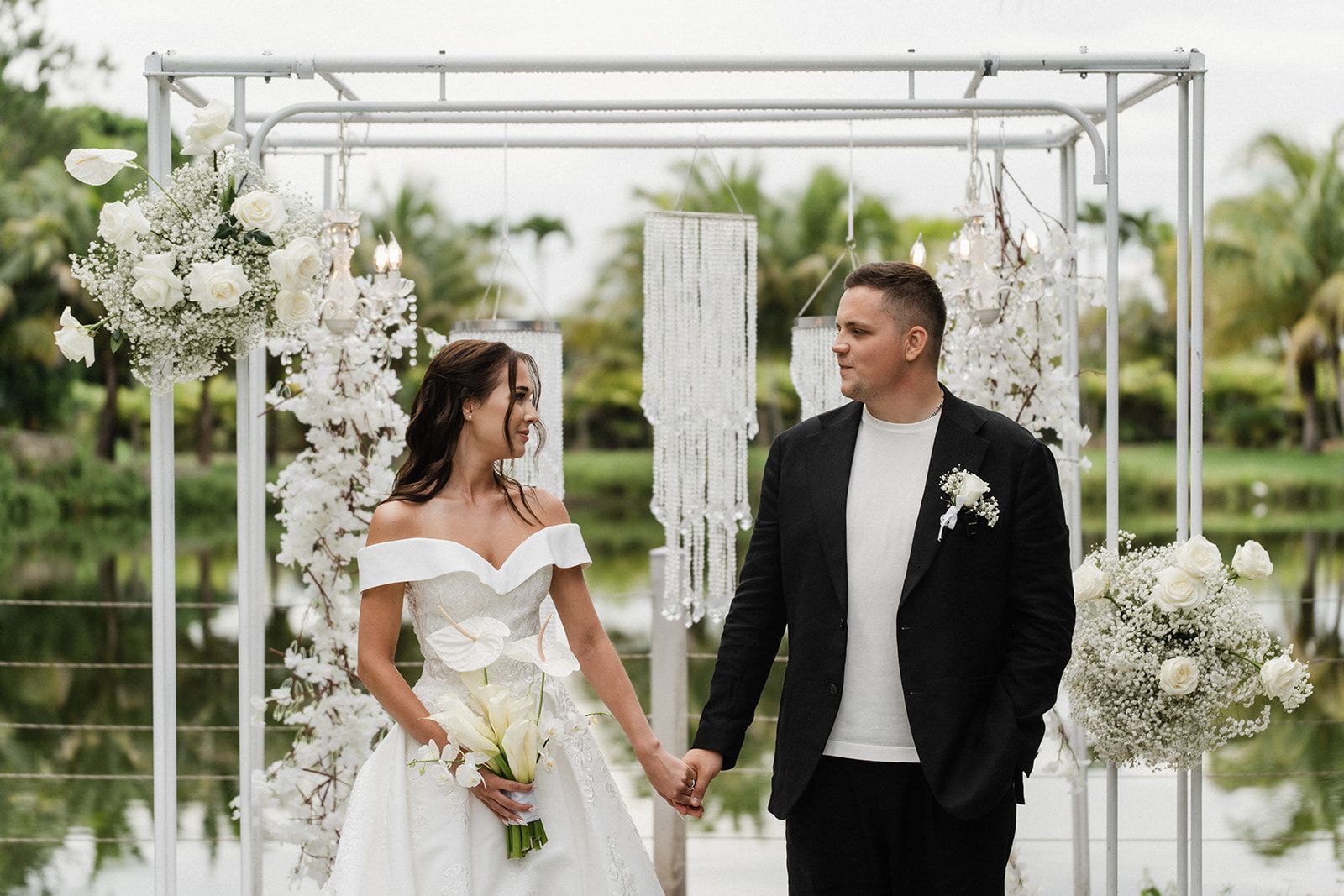 Bride and groom holding hands, smiling at each other under floral archway near a lake.