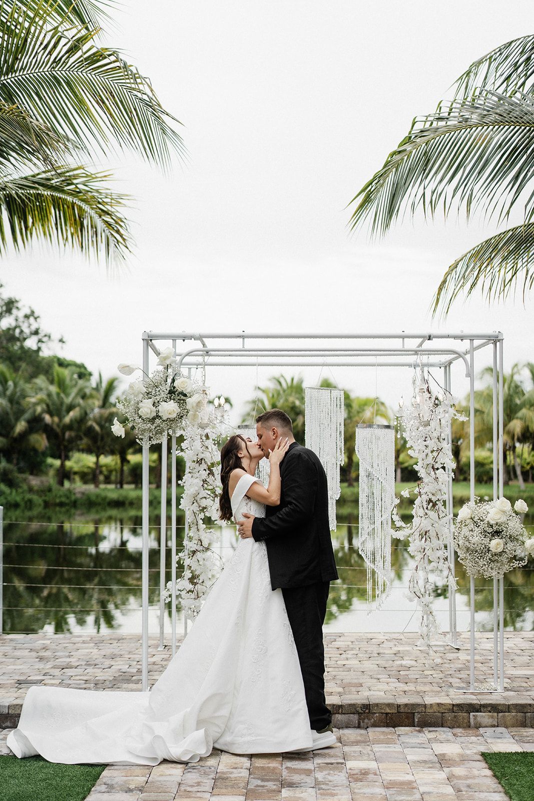 Couple kissing at outdoor wedding ceremony by a pond, white floral arch.