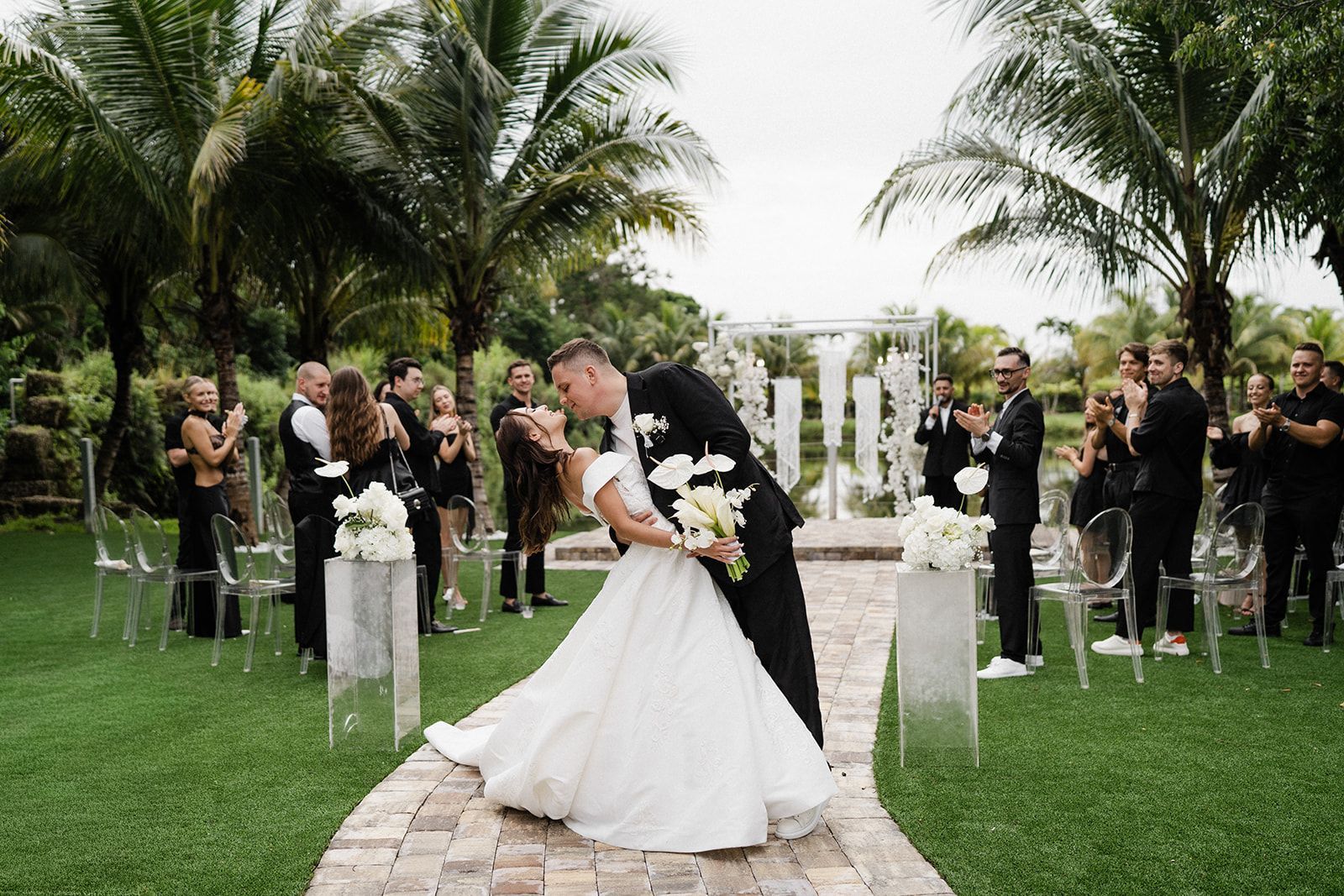Wedding couple kissing on a stone path, surrounded by guests in a lush outdoor setting with palm trees.