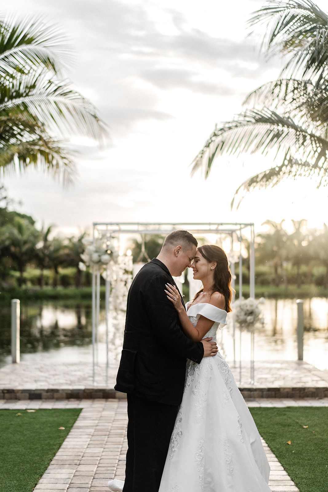 Newlyweds embrace near a lake at sunset. The bride wears a white gown, the groom a black suit.