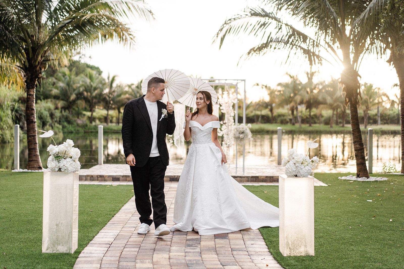 Couple walking, smiling, holding parasols, wedding ceremony. Brick path, floral pillars, water backdrop, palm trees.